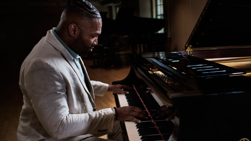 In profile, a Black man in a light, linen suit sits at the piano, his hands on the keys and his eyes are closed reverently. 