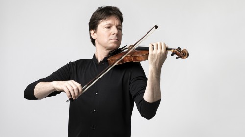 A white man with brown hair plays violin in a black shirt in front of a white background. 