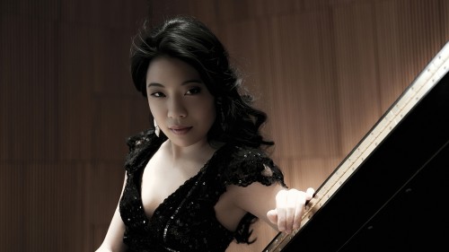 A South Korean woman in a black sparkly dress leans on a grand piano lid, her dark curly hair cascading. 
