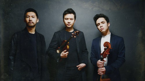 three young men stand in front of a gray portrait background, one holding a violin and one a cello.
