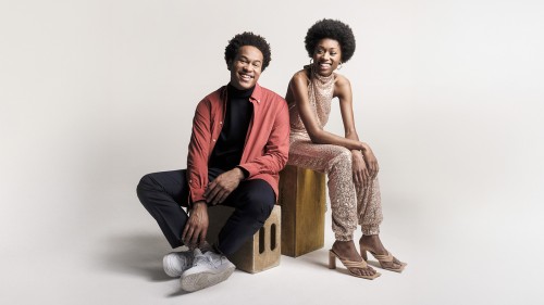 Two young black siblings pose seated on boxes smiling widely, the woman dressed in a glittery jump suit and the man in a red shirt. 