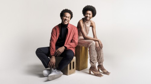 Two young black siblings pose seated on boxes smiling widely, the woman dressed in a glittery jump suit and the man in a red shirt. 