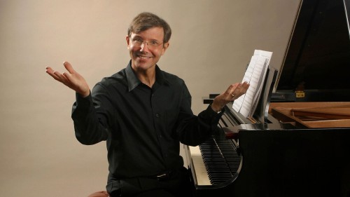 A white man with brown hair and glasses sits at a piano, gesturing as if to say "ta-da!" as he demonstrates a musical concept