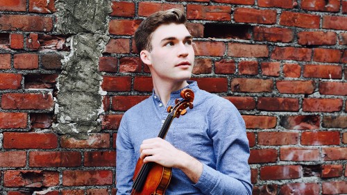 Alexi Kenney a white man in a blue shirt stands in front of a brick wall holding a violin. 