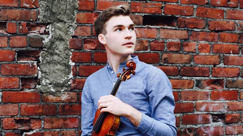 Alexi Kenney a white man in a blue shirt stands in front of a brick wall holding a violin. 