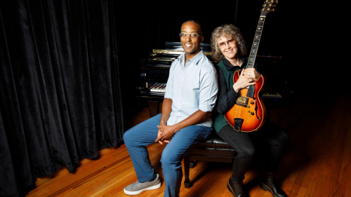 A black man in jeans and a casual buttondown leans against a white woman holding a hollowbody electric guitar. They sit on a bench in front of a grand piano