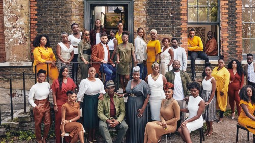 A gospel choir of 30 people poses together on various levels in front of a multi-colored brick building wearing a unifying color palette of white, dark yellow, and shades of terra cotta brown. 