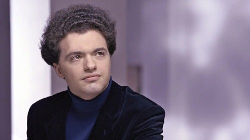 Evengy Kissin, a white man with dark voluminous curly hair in a blue turtleneck and a blazer in front of a purple wall with shadows. 