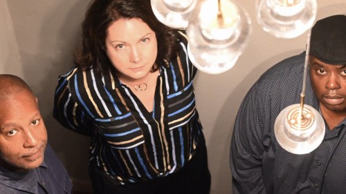 A white woman stands between two black men; the trio looks up with stoic expressions towards a lighting fixture with clear globes. 