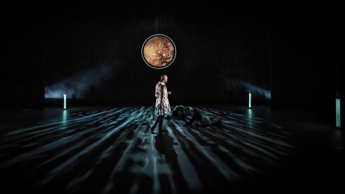 a male dancer stands alone on a dark stage, turned away from us toward two prone dancers and a moon-like illuminated circle