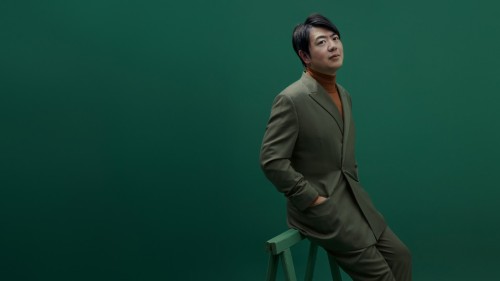 A Chinese man in an olive-green suit and dark hair parted to the side leans on a thin bench, surrounded by a forest green backdrop. 