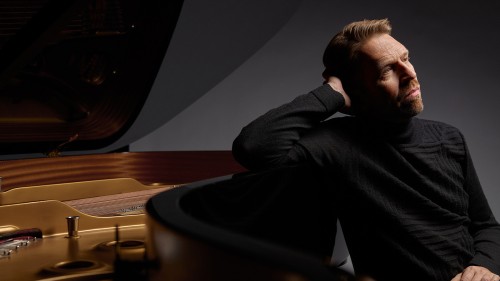 A white man in a black sweater sits next to a grand piano, leaning one arm against the curve of the piano.