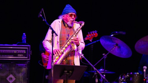 An older Black man, with a white goatee and wearing a knit cap and aviator sunglasses,  plays a tenor saxophone on stage in front of his band.