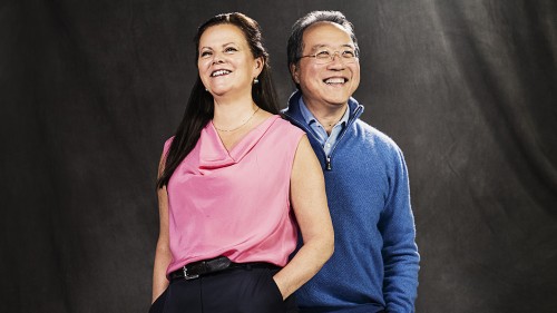 a white woman with long brown hair and a pink top and a gray-haired asian man with glasses and a quarter-zip blue pullover stand with their hands in their pockets, smiling happily. They look like they just shared an inside joke.