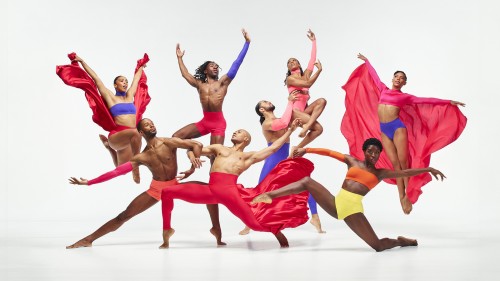 eight dancers, all men in colorful form-fitting pants and the women in colorful bra tops and shorts, leap and pose. A male and a female dancer have billowing red capes.