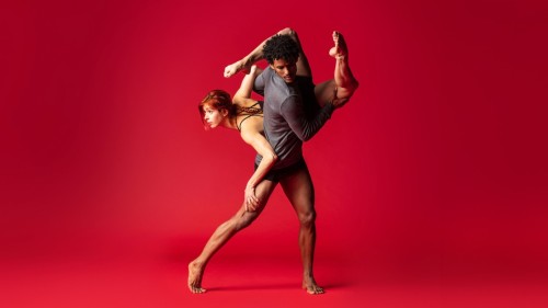 Before a red backdrop, a tall man of twists his torso as he holds a woman with long red hair behind his back in a downward dance lift. The woman’s legs are bent dramatically at the knees as her arms anchor her body to him. 