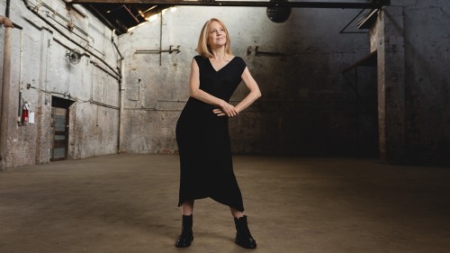 a blonde white woman in a black dress stands with a hand on her hip in an industrial building