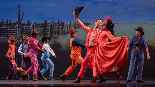 Before a western backdrop, a white male and Black female dancer perform in elaborate coral costumes. While gazing at her, he extends his right arm holding a cowboy hat, and she sweeps her long skirt to the side dramatically. 