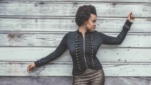 An Ethiopian American woman leans against a wall of grey wood planks looking to the side with her arms outstretched and  wearing a black jacket with gold button details on each side.  