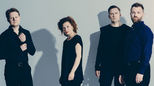 Four Finnish string quartet musicians stand in a line, all wearing dark black and blue clothing, starting solemnly ahead. 