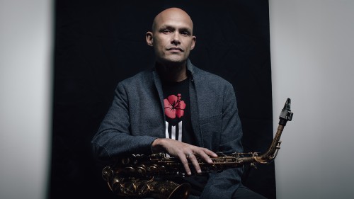 A Puerto Rican man in a gray jacket sits with a saxophone across his lap in front of a black background.