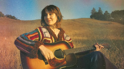 A young white woman, smiling and looking to her right, sits in a field with her guitar. The photo has a vintage effect and evokes late-afternoon "golden hour" sunlight