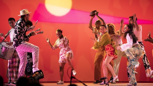 A multiculutral group of dancers clap and play percussion joyfully. They are split into two groups, and a black woman is beginning to dance between the groups as they cheer her on.