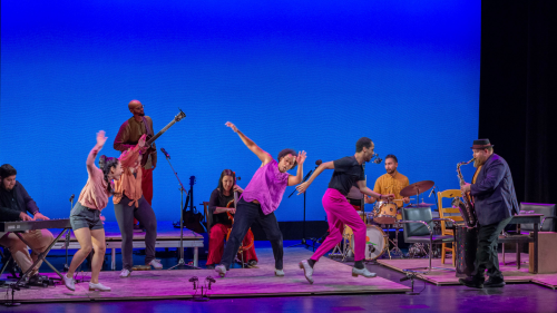 A multicultural group of tap dancers and musicians perform on a series of platforms of varying heights. The staging depitcts the rooms of a house