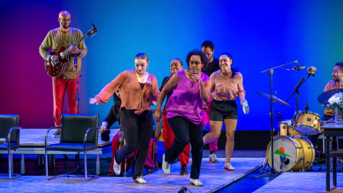 A multicultural group of dancers and musicians clap and tap dance joyfully on a stage set with platforms