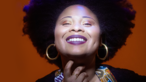 A woman with a large, dark afro smiles with hand under chin wearing large gold hoop earrings, a brightly colored scarf, and purple lipstick as light brightly illuminates her face in front of a dark orange background. 