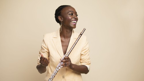 A black woman in a light tan jacket looks to the side, holding her flute in one hand across her body. She smiles warmly.