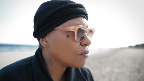 A black woman with a black knit cap, black collared shirt, and sunglasses looks into the distance outdoors. 