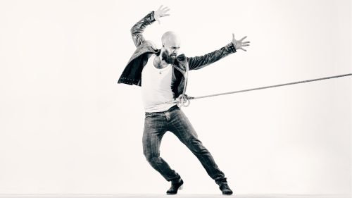 A black and white picture of a bald man with a beard poses with his arms up and reaching to the right side. He leans hard to the left, and a rope around his waist pulls him back towards the center of the frame. 