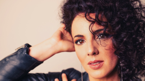 a woman with dark eyes and dark curly hair rests her temple on her hand in a pose evoking 1980s album covers