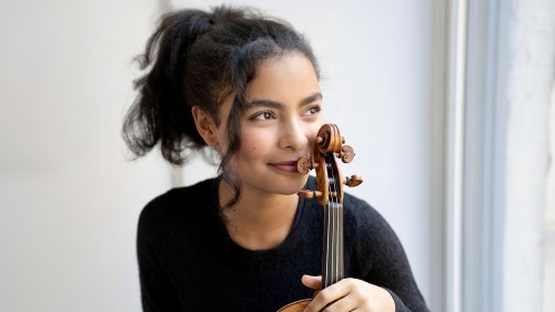 A young woman holds her violin upright wearing a black sweater with her dark curly hair pulled back in a ponytail.