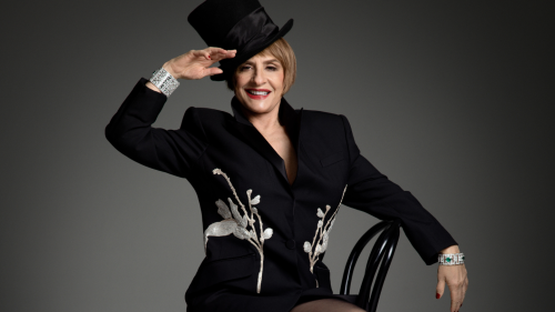 a white woman in her early 70s wears a black skirt suit and lifts a hand to her top hat. she perches on a wooden stool and smiles