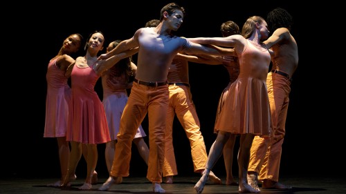 Paul Taylor dancers in a circle gripping arms wearing orange and pink tones. 