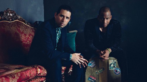 Two Cuban musicians sit in a dark room staring straight ahead, one is on an ornate red couch, one is on a vintage travel suitcase. 