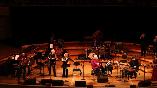 A large group of musicians are spread across a tiered stage with an older man in a red shirt plays the violin on a scooter power chair.