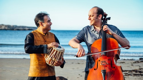 a south asian man holding one small drum and a white man bowing a cello stand on the beach, smiling at each other