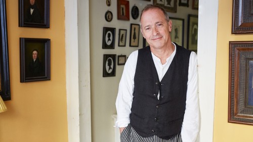 David Sedaris, a white man in his sixties stands in a white shirt with a black vest leaning against a doorframe surrounded by photos on yellow and white walls. 