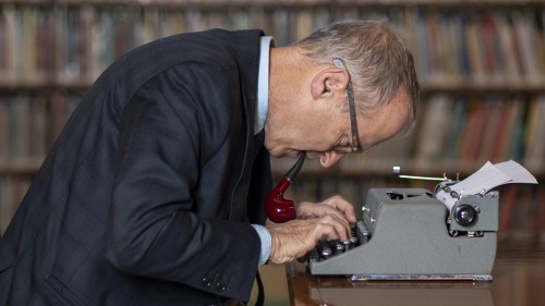 A white man in a jacket and glasses with pipe a dangling out of his mouth leans over a grey typewriter in front of a bookcase. 