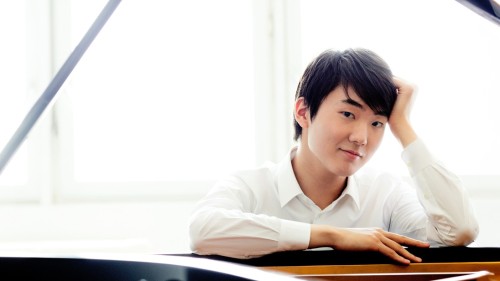 A young, south Korean man in a white collared shirt leans onto the front rim of a grand piano with a slight smile in front of bright windows. 