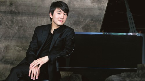 a young east asian man in a dark suit leans on a piano. his elegant hands rest on his lap.