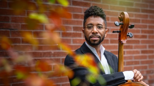 A Black man with gold rim glasses in a dark grey suit stands against a brick wall holding his cello with the strings facing inward while colorful leaves appear out of focus, framing his face.  