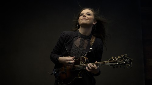 A young white woman with loose brown hair in a black lace top and a blazer, looks up and smiles as she plays her mandolin.