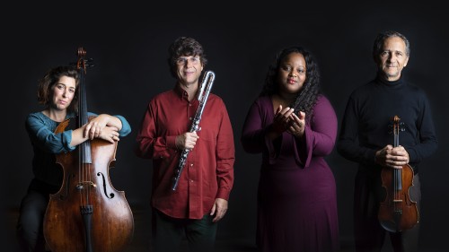 four musicians stand in spotlights - a cellist, a flutist, a vocalist, and a violinist - against a plain dark background
