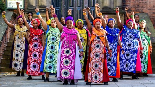 the singers of soweto gospel choir, each wearing a different vibrant color, stand powerfully with fists raised in front of a brownstone building