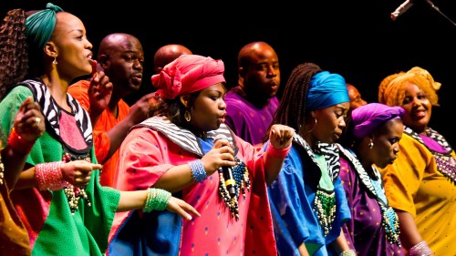 A large choir of African gospel singers perform in brightly colored outfits with smiles on their faces as they sing. 
