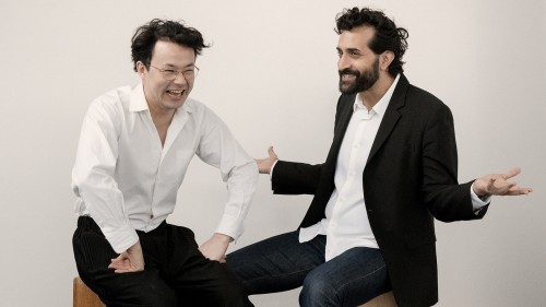 Two young men in a photo studio. The man on the left, with wavy dark hair and glasses, laughs while the man on the right broadly gestures and smiles